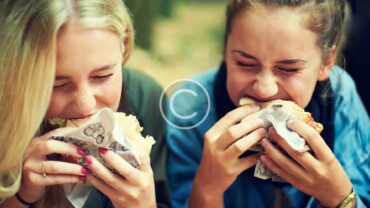 Two girls eating burgers in a park.
